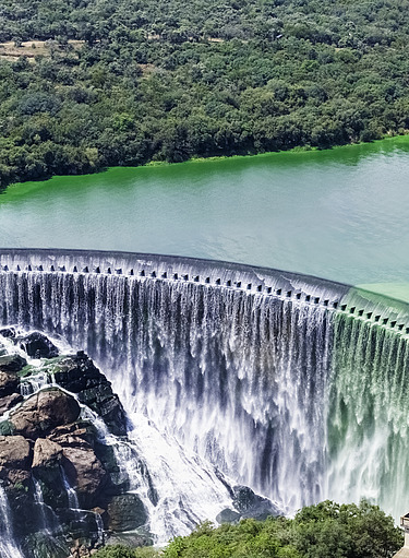 Aerial view of a large curved dam with water flowing over its spillway into a rocky area below. 